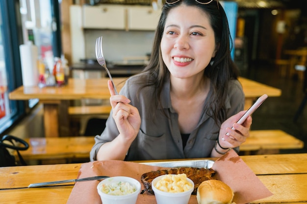 una alegre clienta asiática que sostiene un teléfono y un tenedor está comiendo un rico desayuno en un acogedor restaurante estadounidense. Mujer encantadora de la mañana de vacaciones con una comida abundante y charlando alegremente en la mesa.