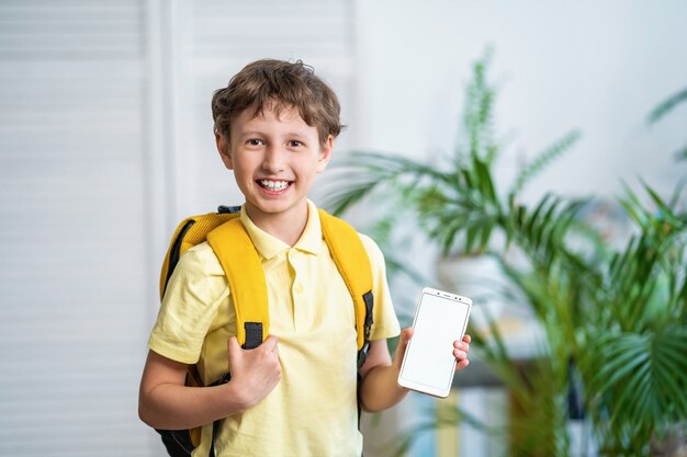 Alegre colegial con una mochila muestra la pantalla blanca del teléfono móvil. E-learning