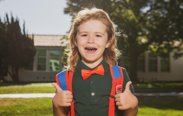 Alegre colegial en uniforme escolar con mochila grande de pie cerca de la escuela de regreso a la escuela k