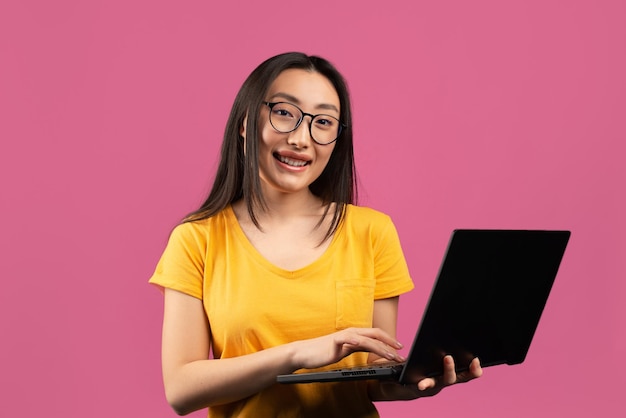 Alegre dama asiática con anteojos sosteniendo la computadora portátil más nueva y sonriendo a la cámara usando un cuaderno estudiando