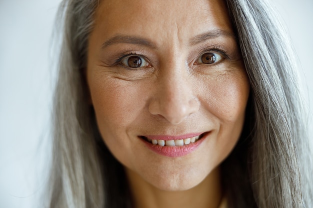 Alegre dama de cabello plateado con maquillaje ligero mira a cámara sobre fondo gris claro en primer plano de estudio. Estilo de vida de belleza madura