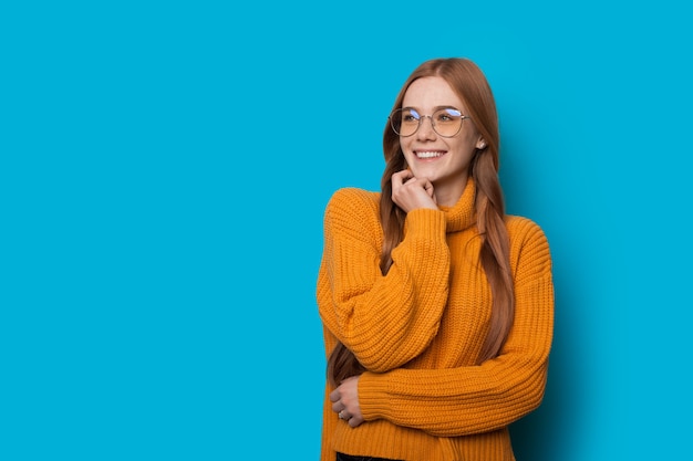 Alegre dama caucásica con cabello rojo y pecas se toca la barbilla mientras posa en una pared azul con espacio libre