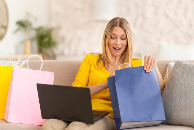 Alegre dama rubia caucásica milenaria adicta a las compras mirando la bolsa de la tienda usando la computadora portátil en el sofá