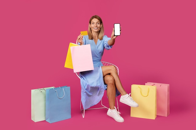 Alegre dama rubia europea de mediana edad con muchos paquetes con compras muestra teléfono inteligente