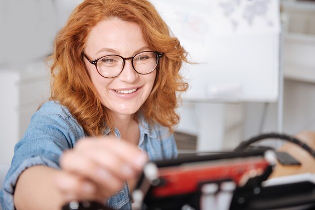 Alegre diseñadora positiva sonriendo y configurando la impresora 3D mientras está de buen humor