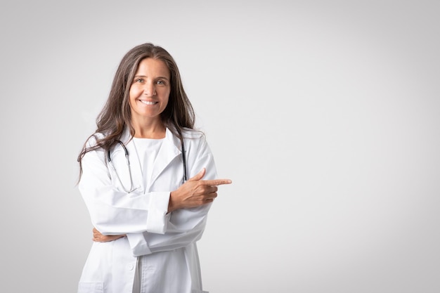 Alegre doctora senior en bata blanca señalando con el dedo el espacio libre aislado en gris claro