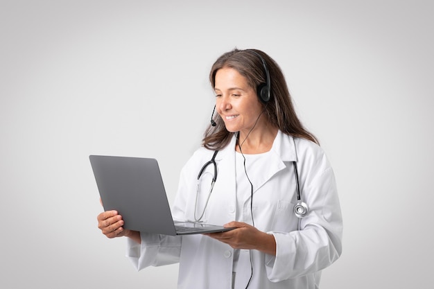 Alegre doctora senior europea en bata blanca con estetoscopio auriculares videollamadas en la computadora portátil