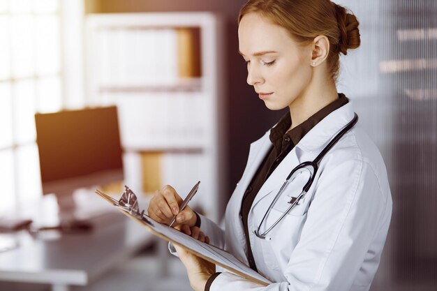 Alegre doctora sonriente usando portapapeles en una clínica soleada. Retrato de una mujer médica amigable en el trabajo. concepto de medicina.