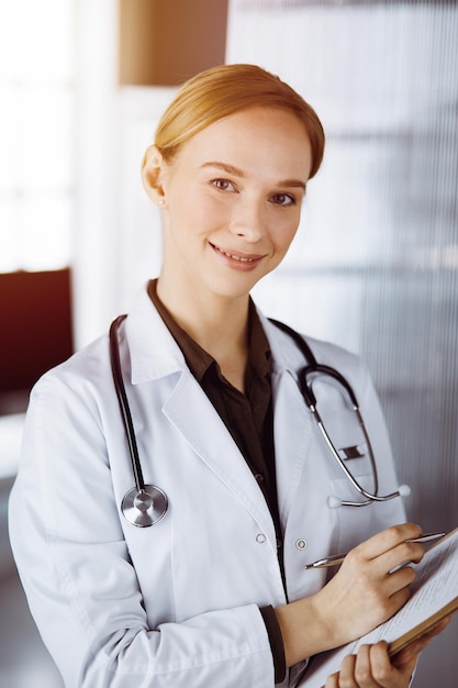 Alegre doctora sonriente usando portapapeles en una clínica soleada. Retrato de una mujer médica amigable en el trabajo. Concepto de medicina.