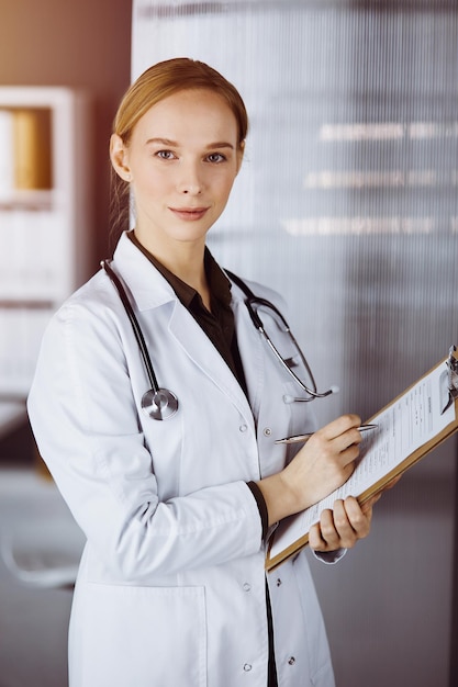 Alegre doctora sonriente usando portapapeles en una clínica soleada. Retrato de una mujer médica amigable en el trabajo. concepto de medicina.