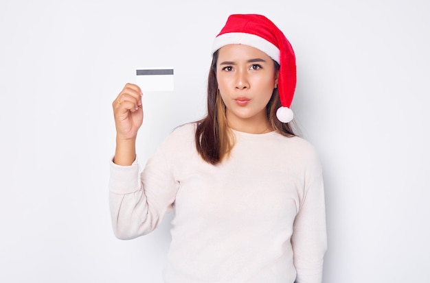 Alegre e animada jovem asiática com um chapéu de Natal mostrando um cartão de crédito branco em branco na câmera isolada no fundo branco