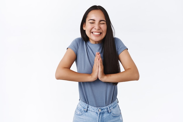 Alegre e animada linda garota asiática acredita que os sonhos se tornam realidade, rezando ou triunfando, junte as mãos em oração, movimento de súplica, feche os olhos e sorria amplamente, alcance o sucesso, desejo realizado