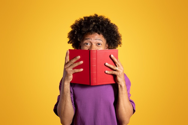 Foto alegre e animado homem afro-americano encaracolado em camiseta roxa parece por trás do livro