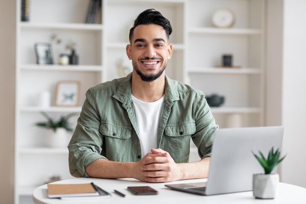 Alegre e atraente empresário árabe milenar com barba no local de trabalho com laptop no escritório em casa