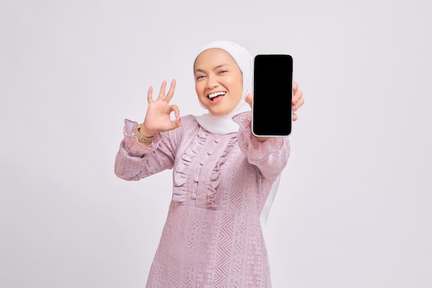 Alegre e bela jovem muçulmana asiática usando hijab e vestido roxo segurando smartphone com tela em branco e gesticulando sinal de ok isolado no fundo branco do estúdio