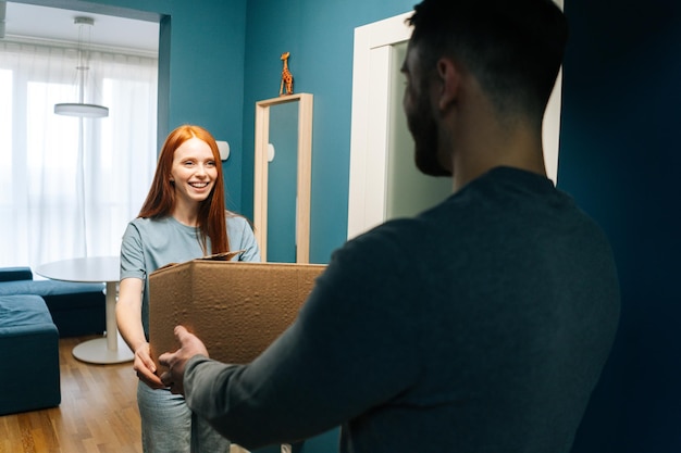 Alegre e bonita jovem recebendo caixa de papelão de entregador irreconhecível na porta do apartamento