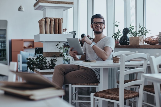 Alegre e bonito. Jovem bonito e moderno segurando um tablet digital e desviando o olhar enquanto está sentado à mesa no café