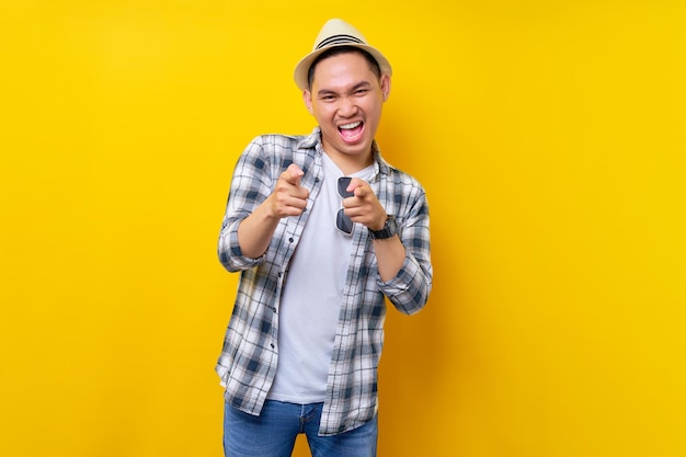 Alegre e bonito jovem étnico asiático de 20 anos usando chapéu de roupas casuais apontando o dedo para a câmera com você enquanto motiva e encoraja isolado no fundo amarelo Conceito de estilo de vida de pessoas