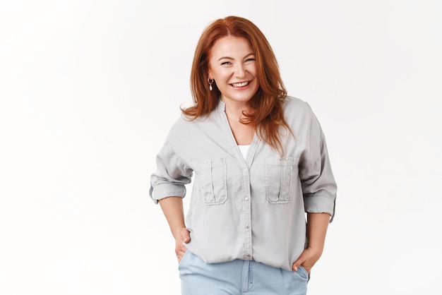 Alegre e carismática ruiva de meia-idade linda mulher sorrindo dentuço feliz de mãos dadas bolsos agradável conversa agradável se sentir alegre relaxado após bons fins de semana de férias fundo branco