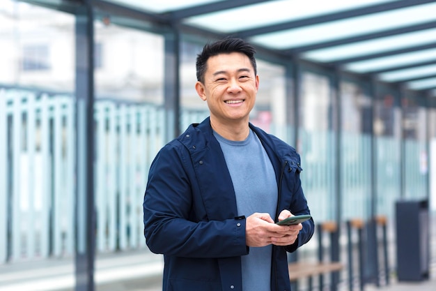 Alegre e feliz asiático usando telefone sorrindo e olhando para a câmera perto do empresário do aeroporto em viagem turística