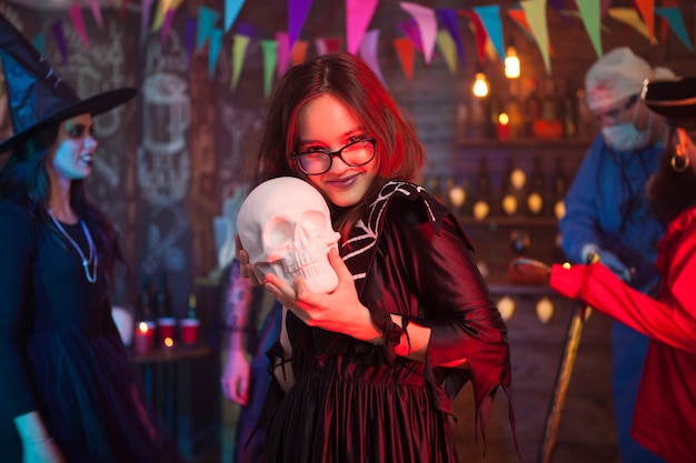 Alegre e linda garota segurando um crânio humano para a celebração do halloween. Garota com fantasia de bruxa.