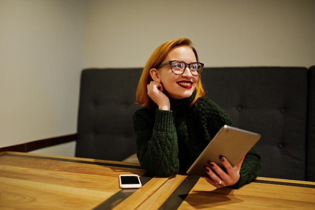 Alegre e linda mulher ruiva de óculos usa suéter de lã verde quente usando seu tablet touchpad enquanto está sentado no local de trabalho no café