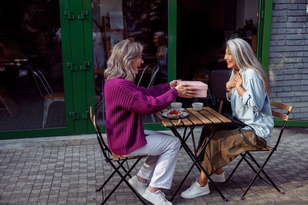 Alegre e madura amiga dando presentes para uma mulher asiática surpresa na mesa de madeira