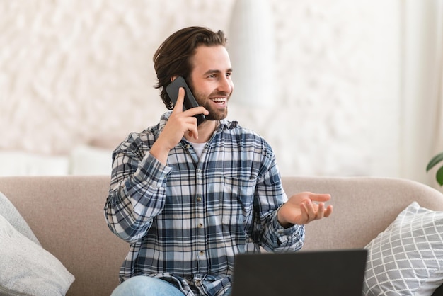 Alegre e ocupado jovem gerente masculino caucasiano sentado no sofá com chamadas de computador por telefone fala com o cliente