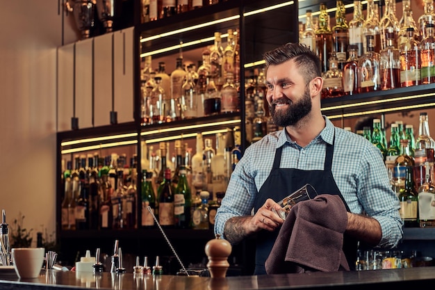 Un alegre y elegante barman brutal está limpiando un vaso con un paño en el fondo del mostrador del bar.