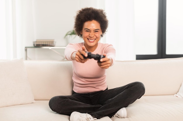 Alegre y emocionada joven mujer de raza mixta sentada en un sofá con joystick jugando juegos de computadora en la sala de estar