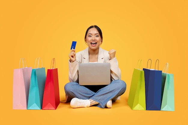 Alegre y emocionada señora china del milenio con bolsas de compras tarjeta de crédito portátil disfrutar de la venta y ganar