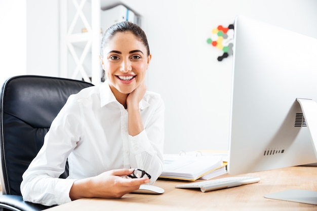 Alegre empresaria linda sentada en el lugar de trabajo usando la computadora en la oficina