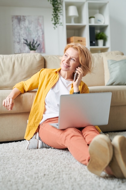 Alegre empresária loira madura com laptop e smartphone, consultando clientes enquanto trabalha remotamente em casa