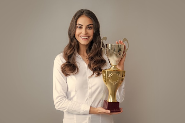 Alegre empresaria sosteniendo el trofeo de pie posando sobre fondo gris