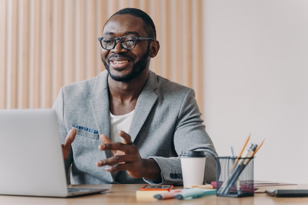 Alegre empresário afro-americano usando óculos e blazer estiloso fazendo videochamada no laptop