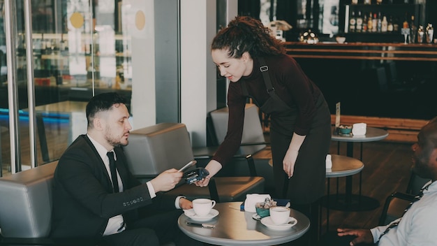 Alegre empresário caucasiano sentado com seu parceiro afro-americano e pagando a conta on-line usando seu smartphone no café moderno jovem garçonete usando terminal de dinheiro