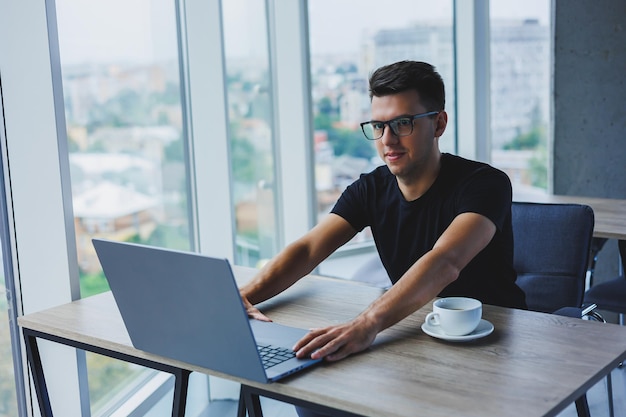 Alegre empresário de óculos se senta em sua mesa trabalhando com um laptop no escritório Um empresário se senta e trabalha em um laptop e bebe café Começando o dia no escritório