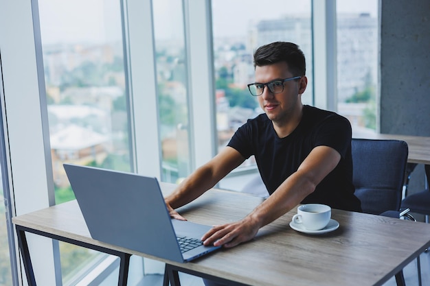 Alegre empresário de óculos se senta em sua mesa trabalhando com um laptop no escritório um empresário se senta e trabalha em um laptop e bebe café começando o dia no escritório