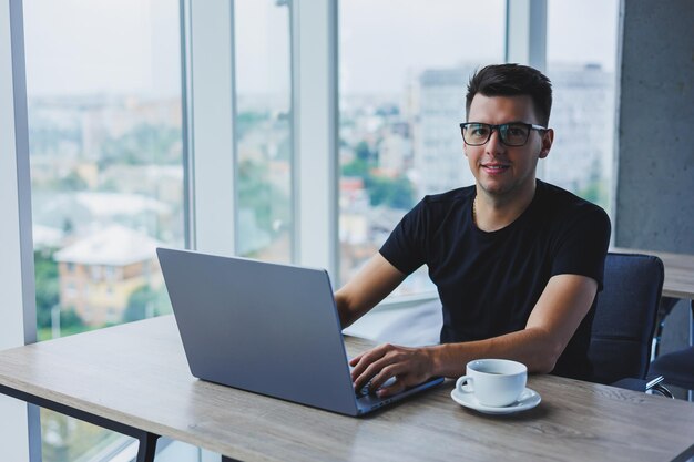 Alegre empresário de óculos se senta em sua mesa trabalhando com um laptop no escritório Um empresário se senta e trabalha em um laptop e bebe café Começando o dia no escritório