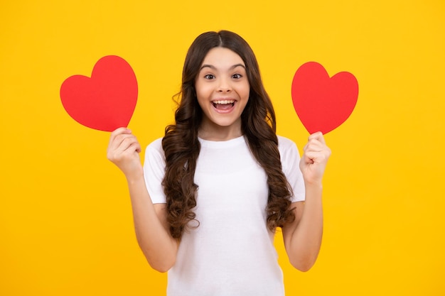 Alegre, encantadora y romántica jovencita sostenga el símbolo del corazón rojo del amor por el día de San Valentín aislado en un fondo amarillo. Adolescente feliz, emociones positivas y sonrientes de la jovencita.