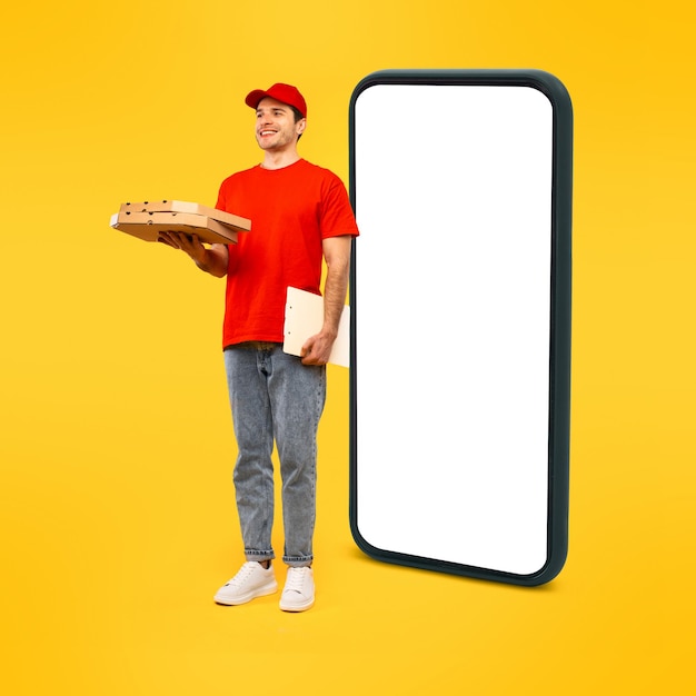 Alegre entregador segurando caixas de pizza em pé perto de fundo amarelo de smartphone