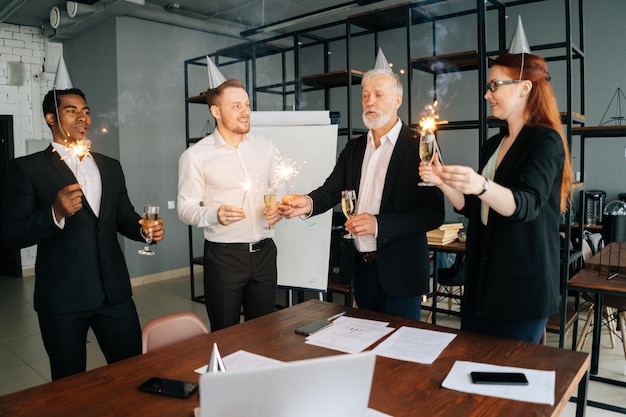 Alegre equipo de negocios de compañeros de trabajo multiétnicos sosteniendo luces de Bengala tintineando copas de champán