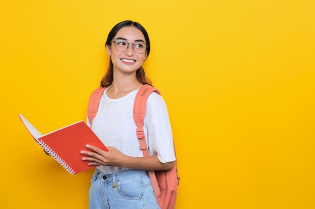 Alegre estudante muito jovem vestindo mochila e óculos segurando livros olhando para longe no espaço em branco isolado em fundo amarelo