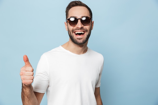 Alegre excitado hombre vestido con camiseta en blanco que se encuentran aisladas sobre la pared azul, mostrando los pulgares para arriba