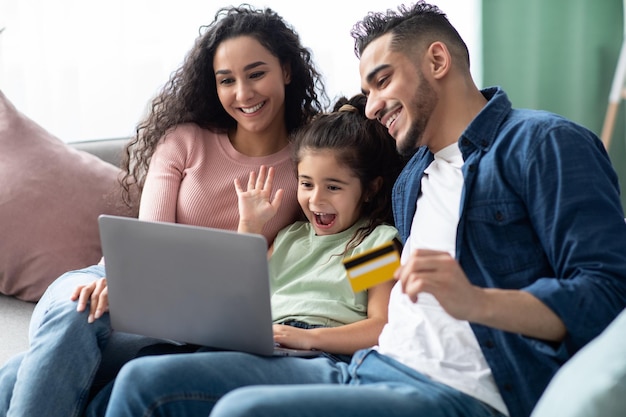 Alegre família árabe de três com laptop e cartão de crédito fazendo compras on-line enquanto relaxa no sofá em casa, pais felizes do Oriente Médio e filhinha aproveitando a compra da Internet