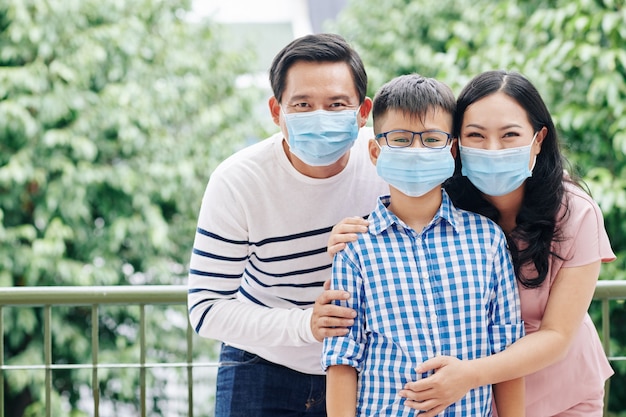 Alegre familia asiática de tres en máscaras médicas de pie al aire libre y mirando