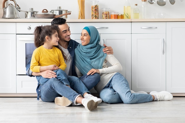 Alegre família islâmica de três relaxando no chão na cozinha juntos, feliz mãe muçulmana em hijab, seu marido árabe e filhinha em casa, abraçando e rindo, espaço para texto