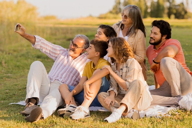 Alegre familia multigeneracional europea disfruta de un picnic en cuadros escoceses en el parque tomando selfie en el teléfono