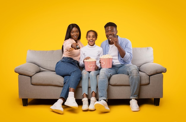 Alegre família negra de três sentado no sofá e assistindo filme comendo pipoca do balde isolado na parede amarela