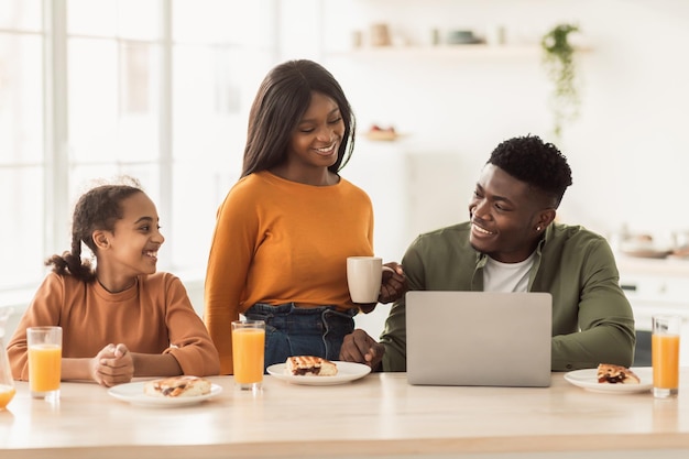 Alegre familia negra usando laptop almorzando sentado en la cocina